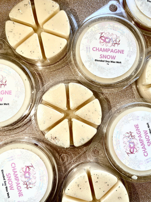 A collection of hand-poured soy wax tarts labeled "Champagne Snow Wax Melt" by The Soap Gals arranged on a surface. Each package contains six triangular pieces, accented with biodegradable glitter.