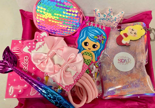 Assortment of colorful children's accessories including a sparkly sequin purse, mermaid notebook, bow hairband, and decorative items on a pink background from The Soap Gal x Girls Treat Box - Ready To Go Gift Box.