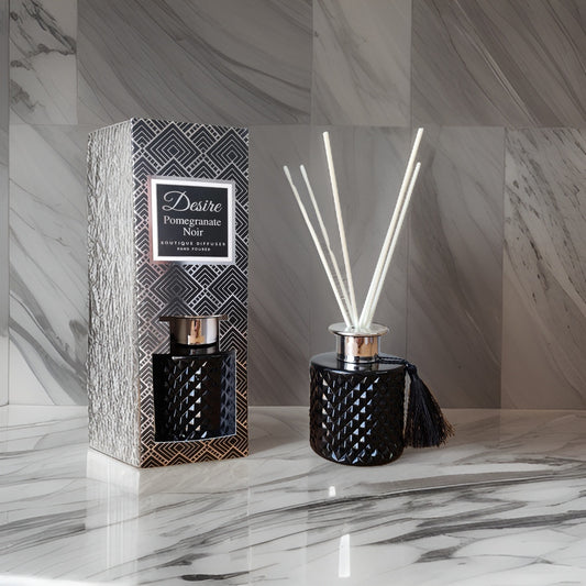 A Pomegranate Noir Reed Diffuser by The Soap Gal x with a black, patterned container next to its packaging on a marble surface.