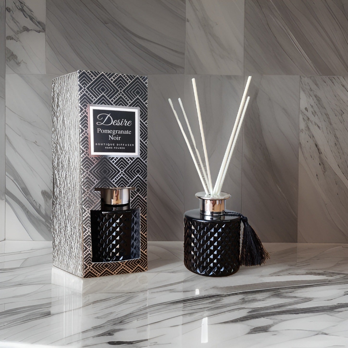 A Pomegranate Noir Reed Diffuser by The Soap Gal x with a black, patterned container next to its packaging on a marble surface.