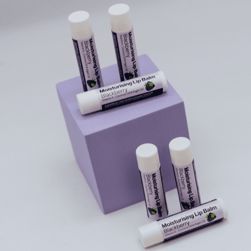 Four tubes of blackberry and thyme handmade Moisturising Lip Balm by The Soap Gal x displayed on a purple stand.