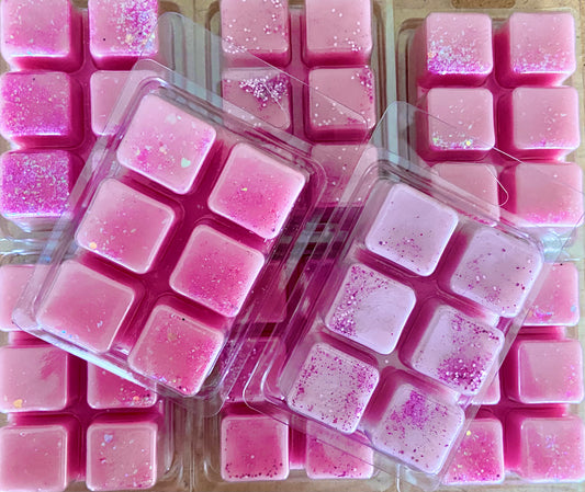 Pink wax melts, some in clamshell packaging and others arranged outside, with sparkling glitter accents on top, laid out in a visually appealing manner. These Soap & Glorious Wax Melts by The Soap Gal x promise a long-lasting fragrance that will delight your senses.