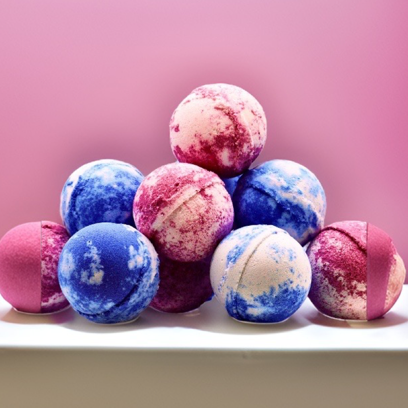 A collection of round, multicolored Random Scented Jumbo Bath Bombs displayed on a shelf against a pastel pink background by The Soap Gal.