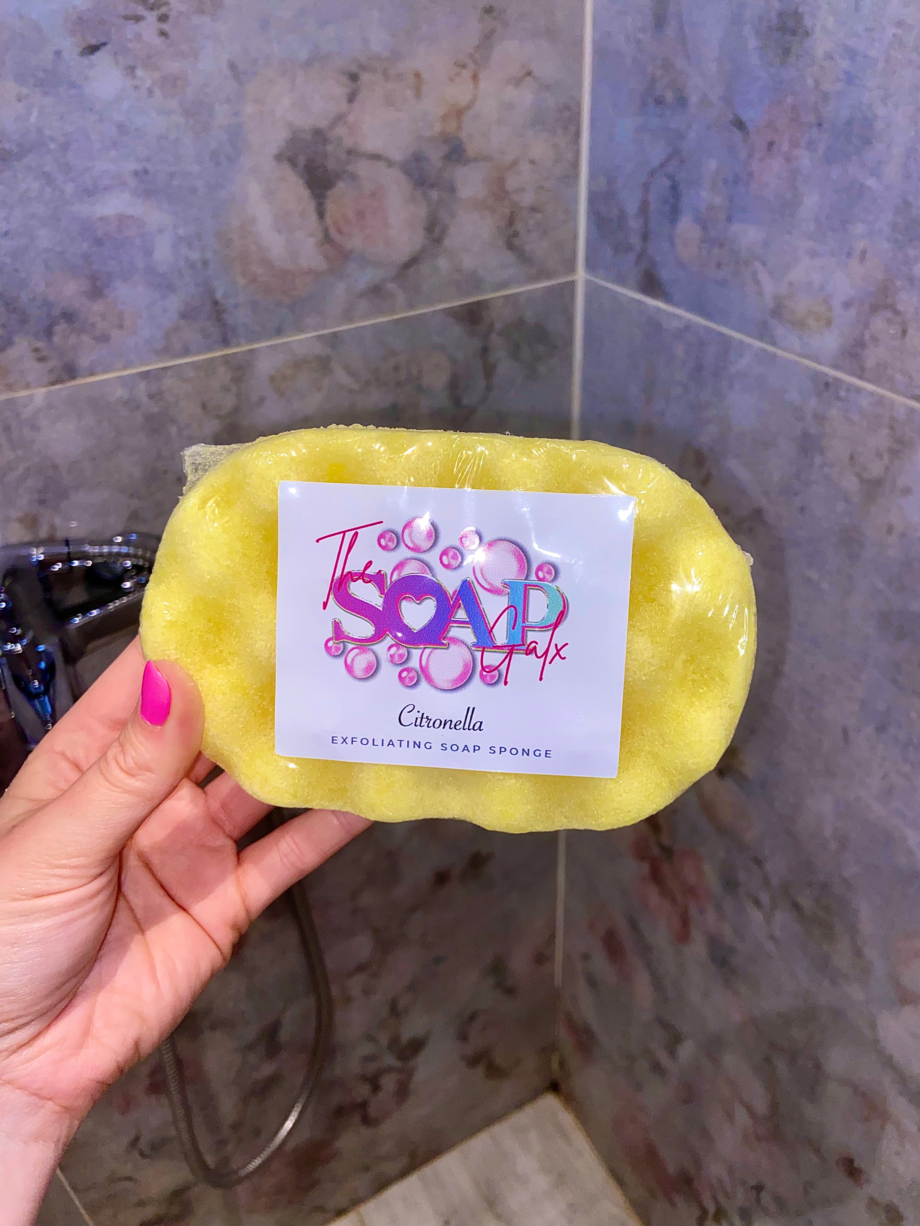 A hand holding a yellow Citronella Soap Sponge by The Soap Gals labeled "tsóraíocht soap co. citronella" against a tiled shower background, offering natural protection.