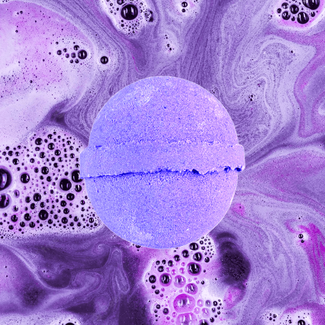 A purple Aliens Bath Bomb from The Soap Gals dissolves, creating swirling patterns with froth and beads of color in water, releasing a fragrant Jasmine Sambac aroma.