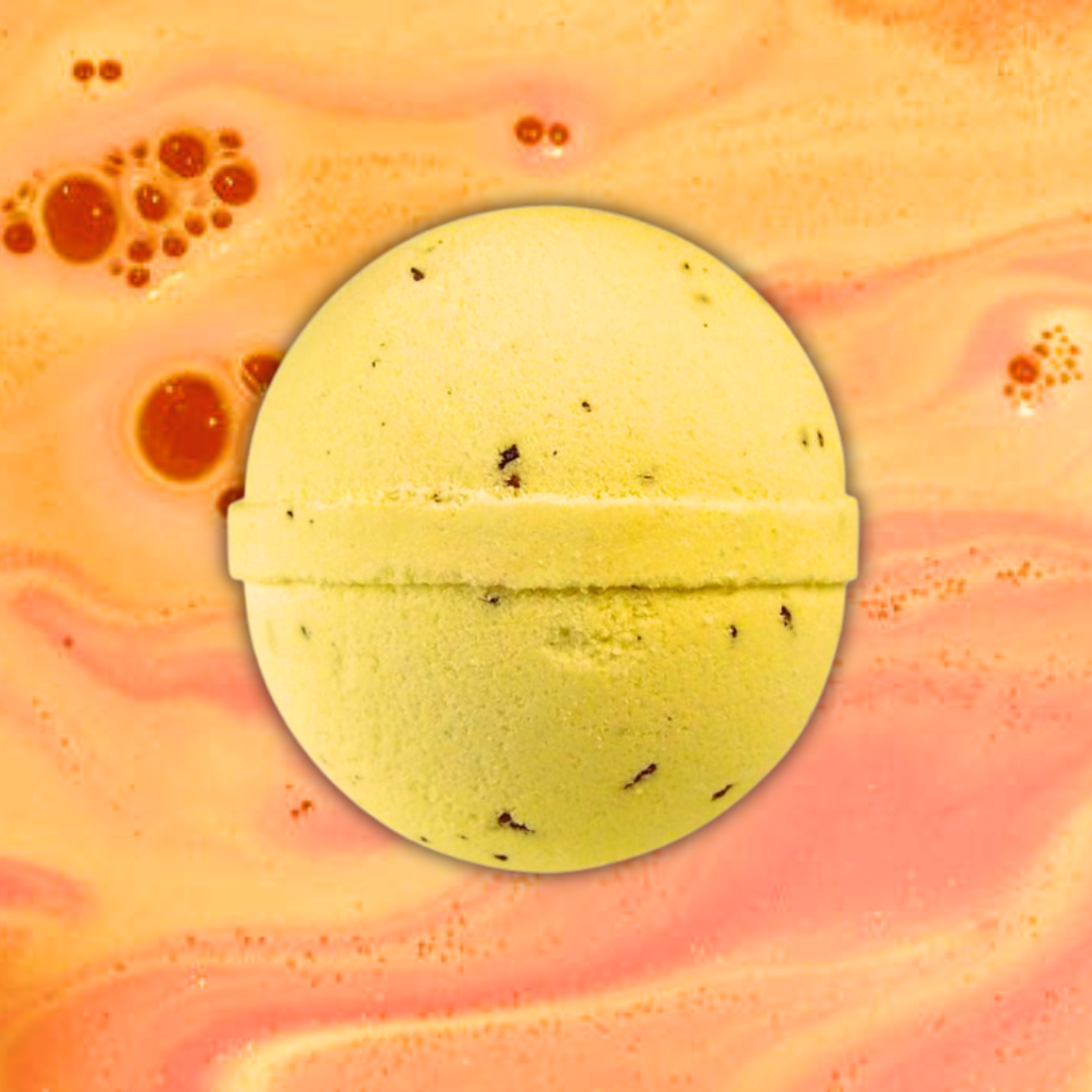 A yellow Toffee Fudge Bath Bomb with black specks centered on a swirling pink and orange pastel background by The Soap Gals.