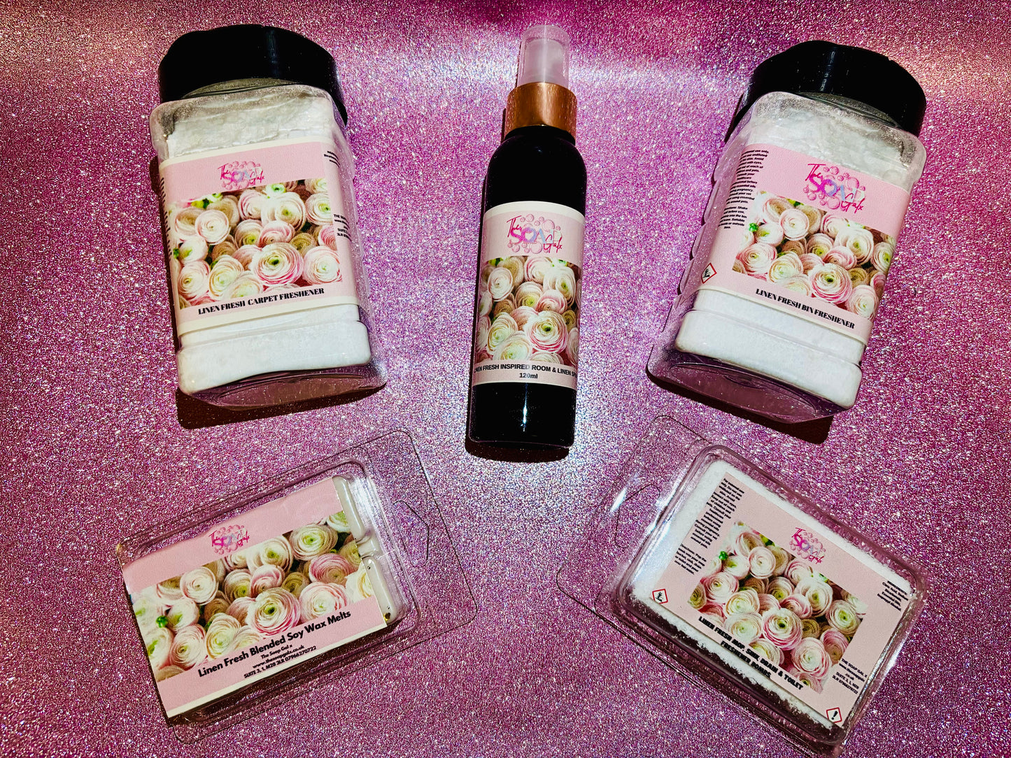 Cosmetic products with floral labels displayed on a glittery pink surface now include The Soap Gals' luxury perfumes.