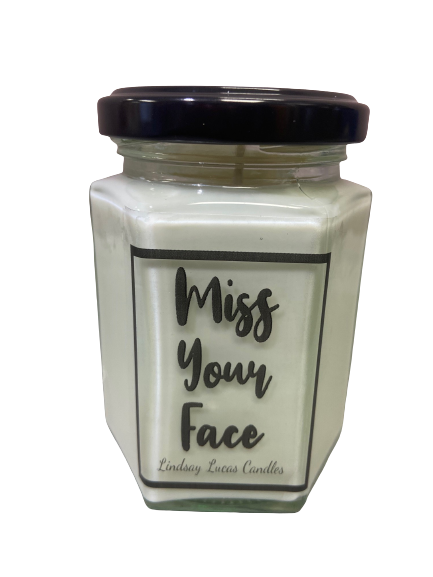 A hexagonal glass jar candle with a black lid, labeled "Miss Your Face Candle" by The Soap Gal x. The white wax radiates the longing and affection of a heartwarming gift.